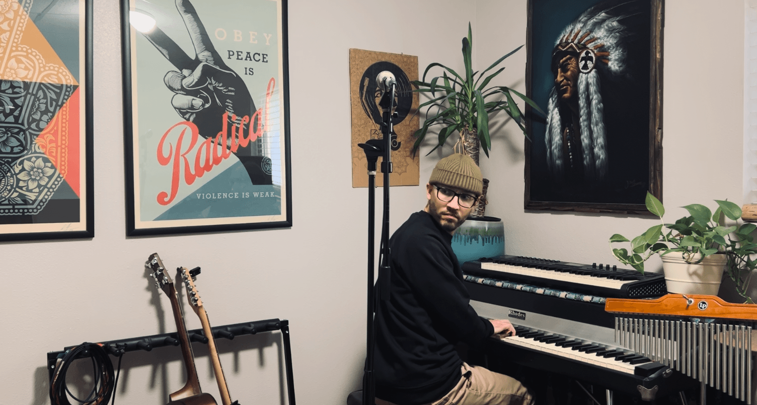 Person wearing glasses and a hat sitting at a piano in a room with framed posters and plants.