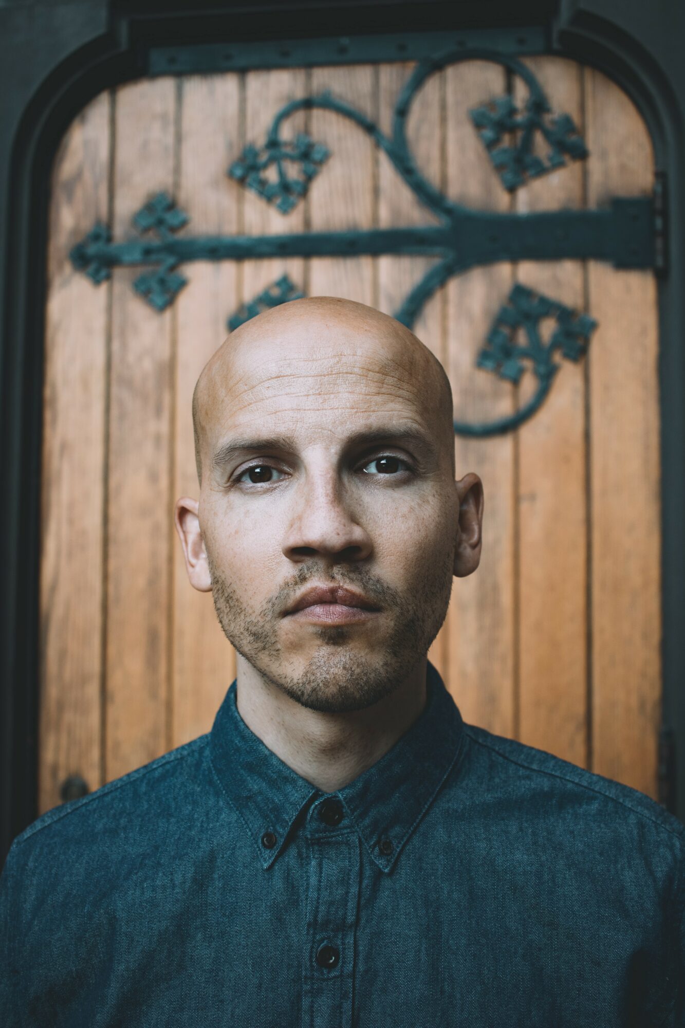 Portrait of a bald man with a serious expression, wearing a dark button-up shirt, standing in front of a wooden door with decorative ironwork.