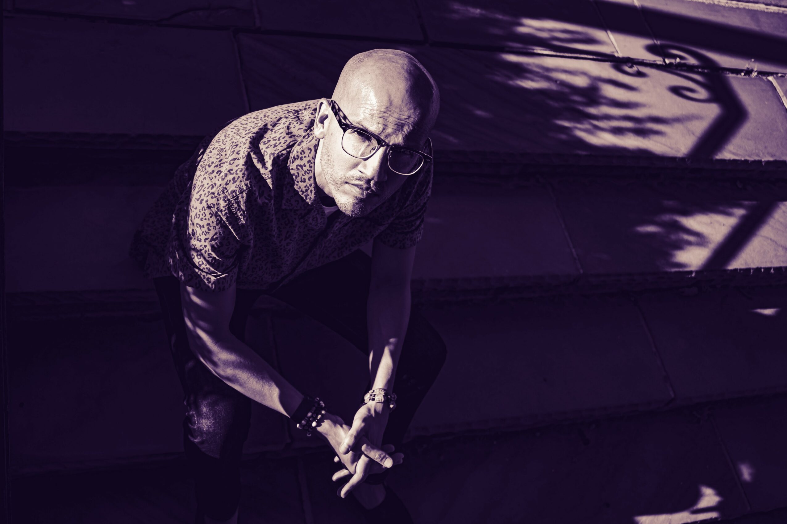 A man with glasses and a patterned shirt crouches on stairs, with shadows cast on the wall behind him.
