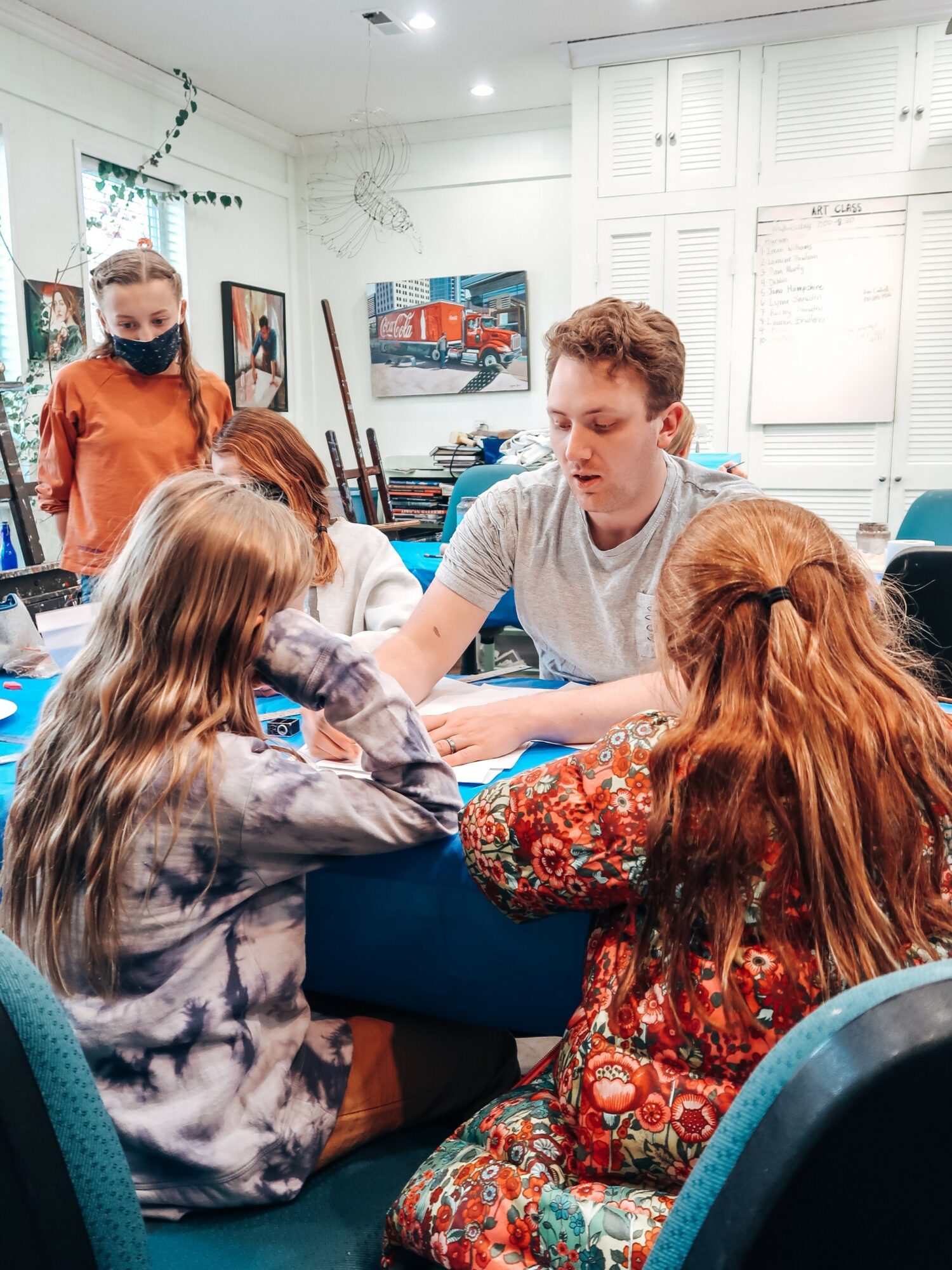 Young man sitting at a table with children, engaging in an activity in a classroom with artwork on walls.