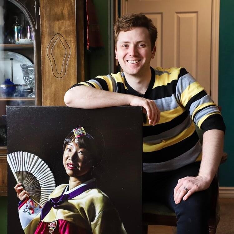 Young man smiling, sitting in a room with a wooden cabinet and a door in the background.