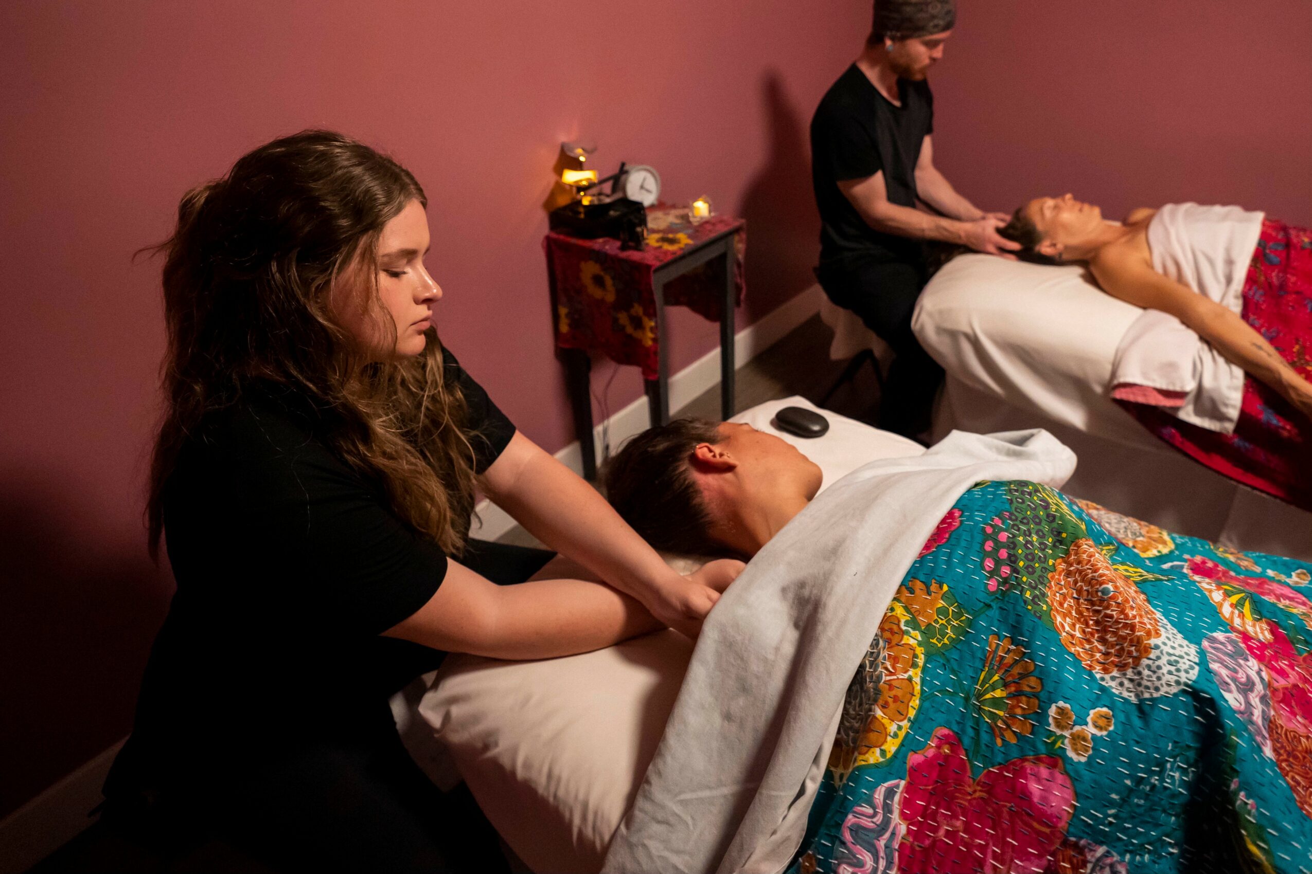 Two massage therapists work on clients lying on massage tables in a room with pink walls, one with a colorful blanket.