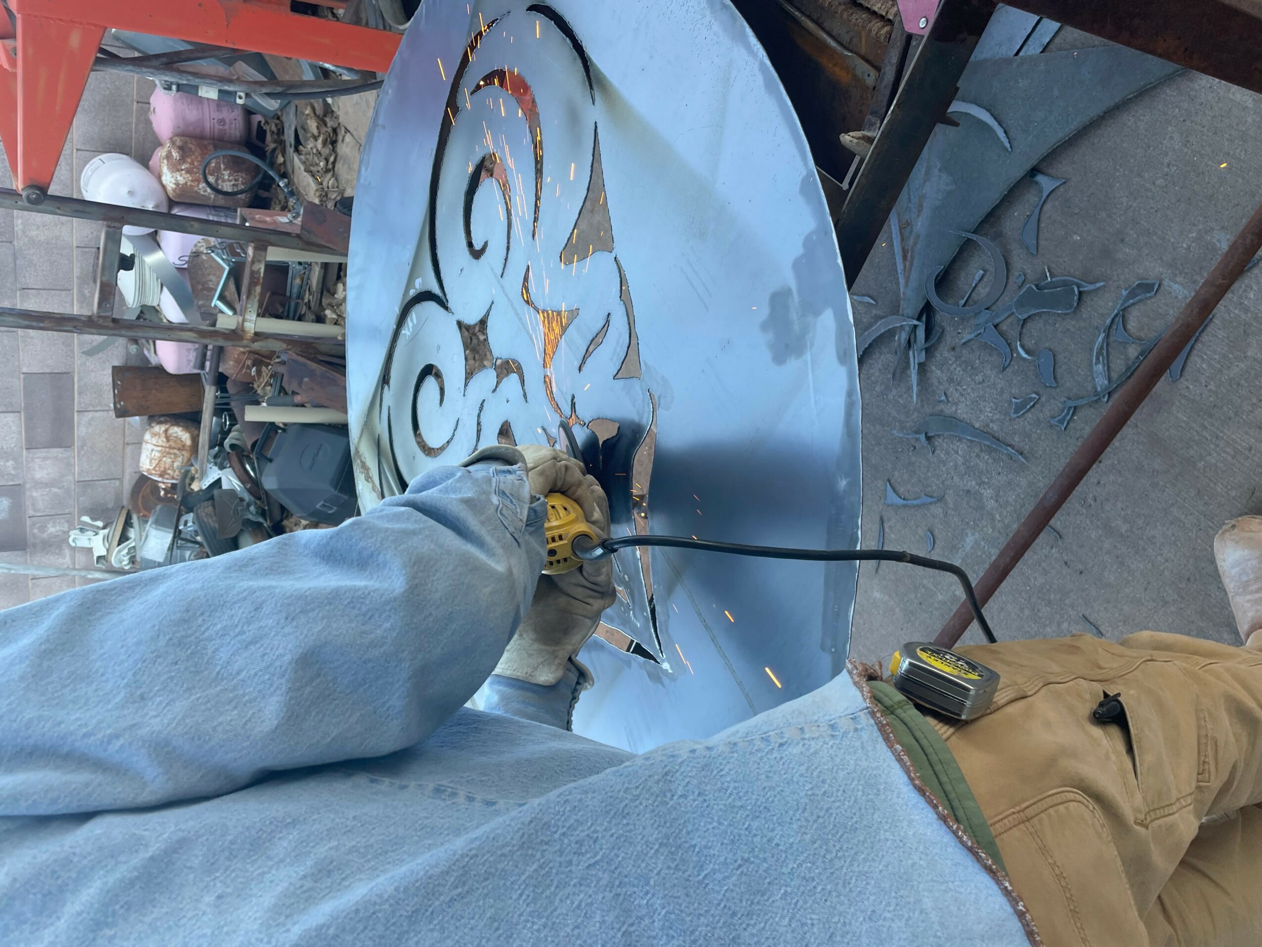 Person using a power tool on a round metal surface with graffiti, in an outdoor workshop setting.