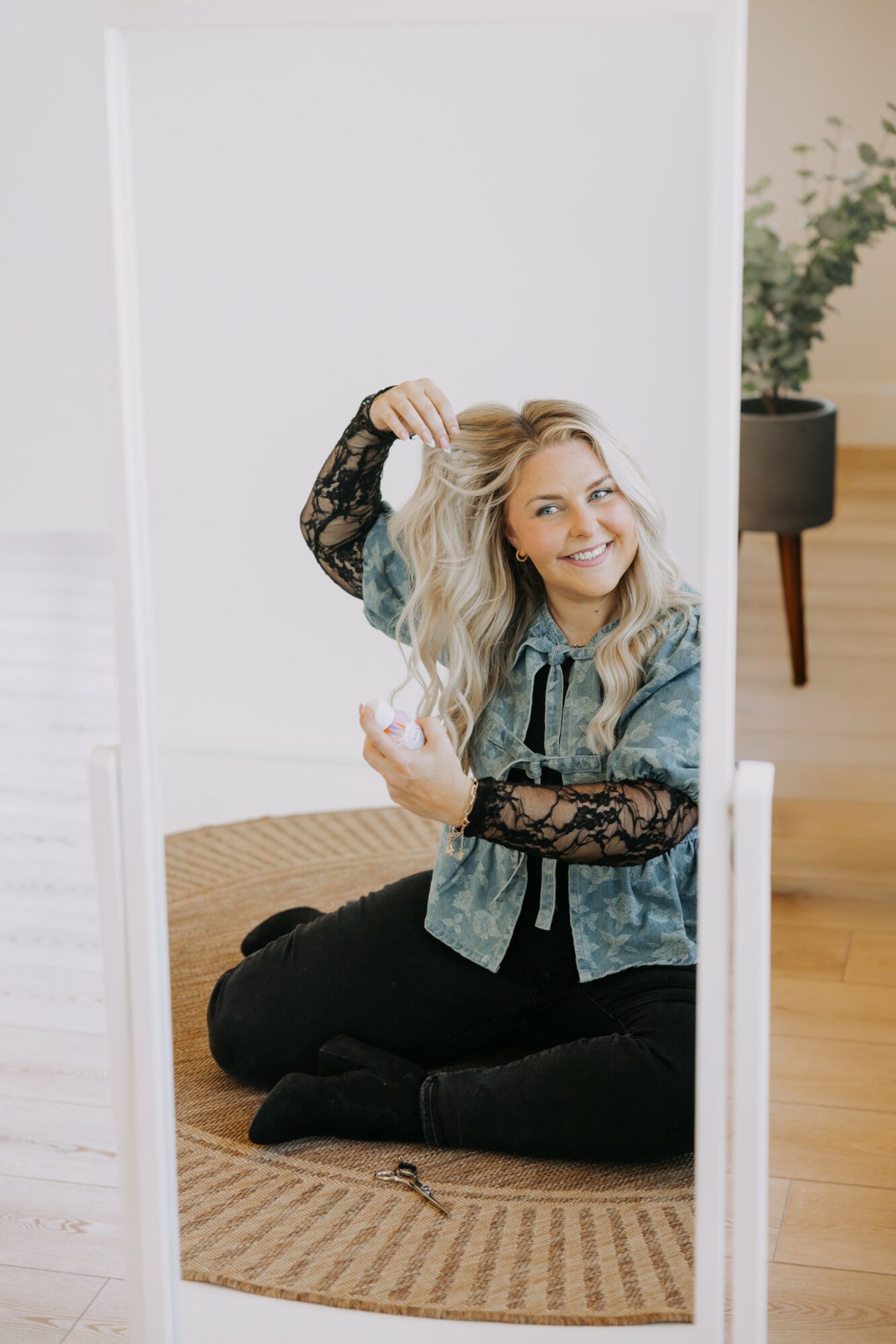 Young woman with blonde hair sitting on a rug, smiling, holding a strand of hair, in front of a mirror.