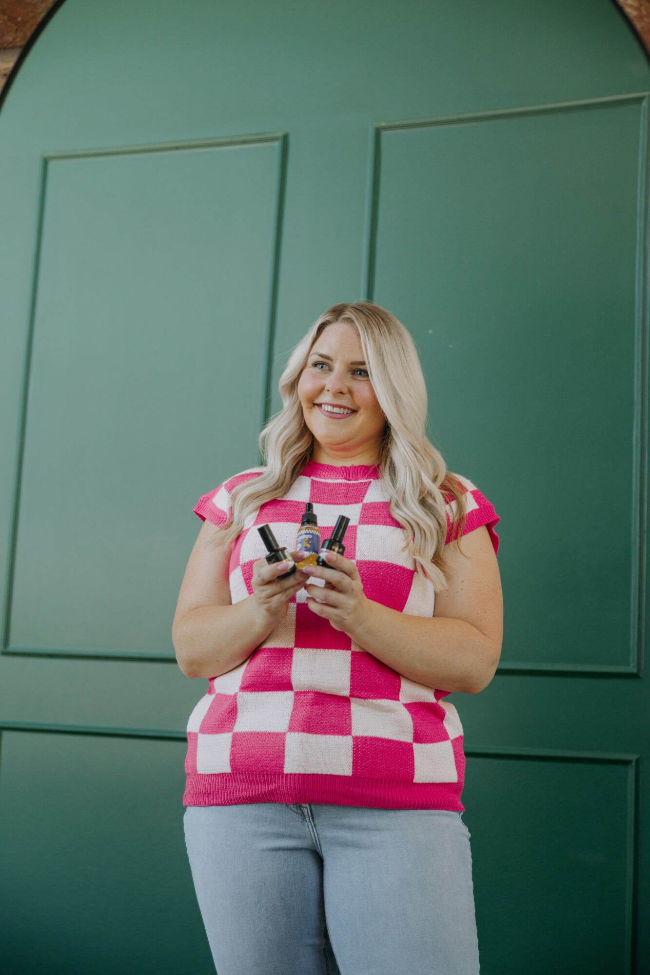 Woman with long blonde hair smiling, holding a smartphone, standing in front of a green wall with rectangular panels.