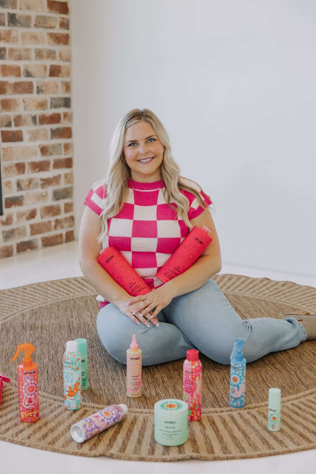 Woman sitting on a rug with colorful bottles and containers around her, smiling, with a brick wall and white background.