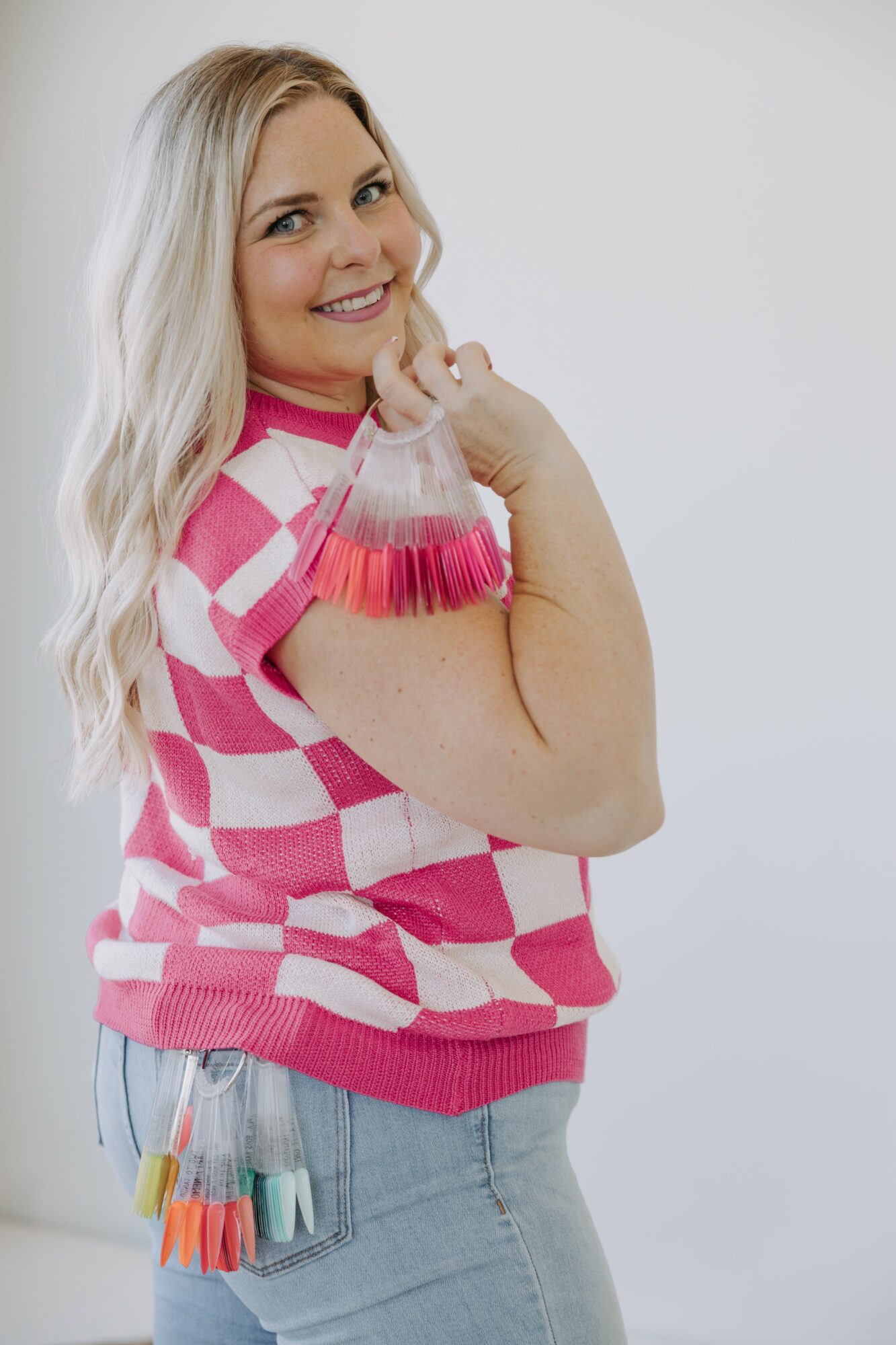 Smiling woman with long blonde hair wearing a pink and white checkered sweater, holding paint swatches, standing sideways.