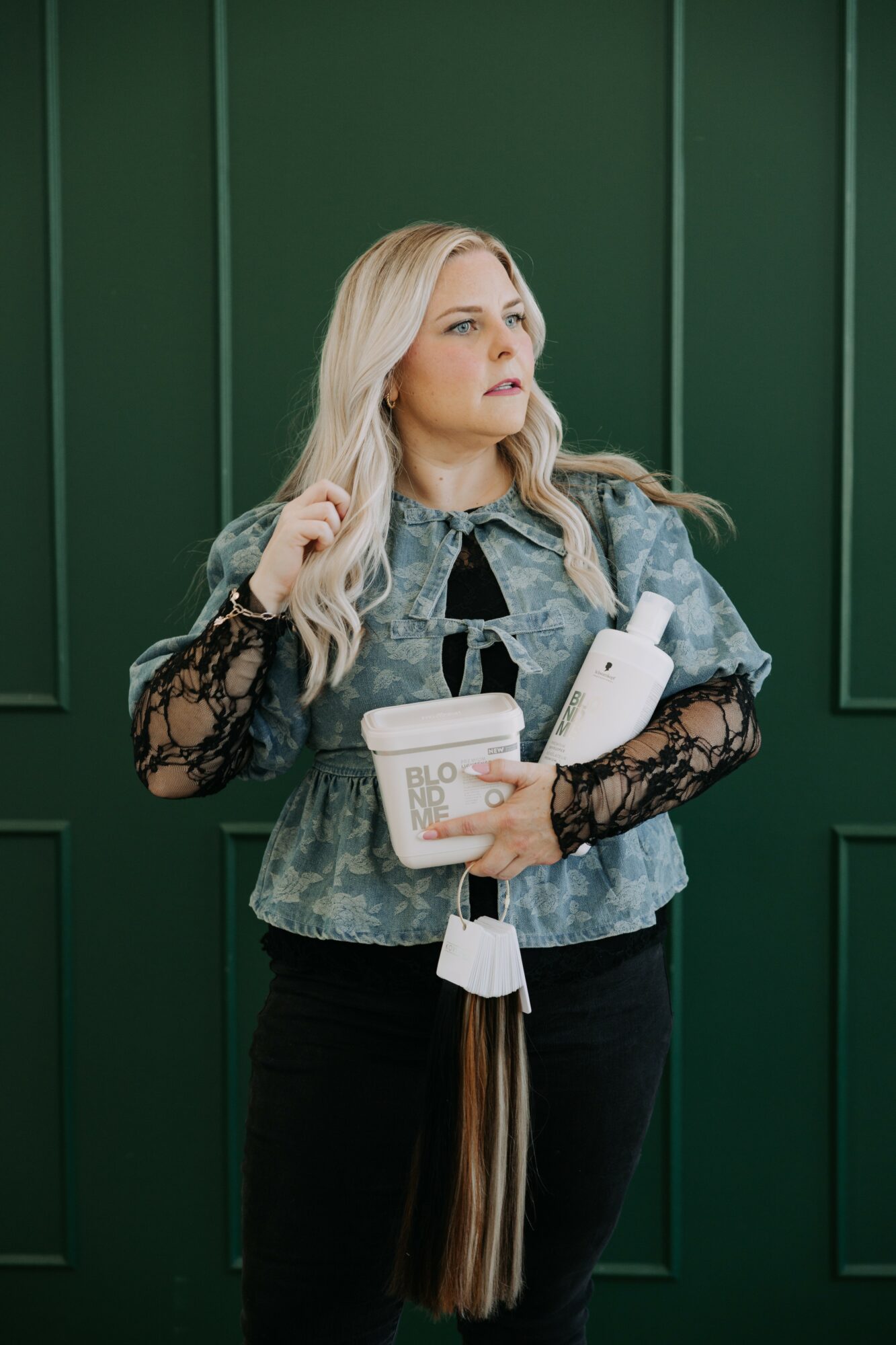 Woman with long blonde hair holding a book and a shopping bag, standing against a green wall.