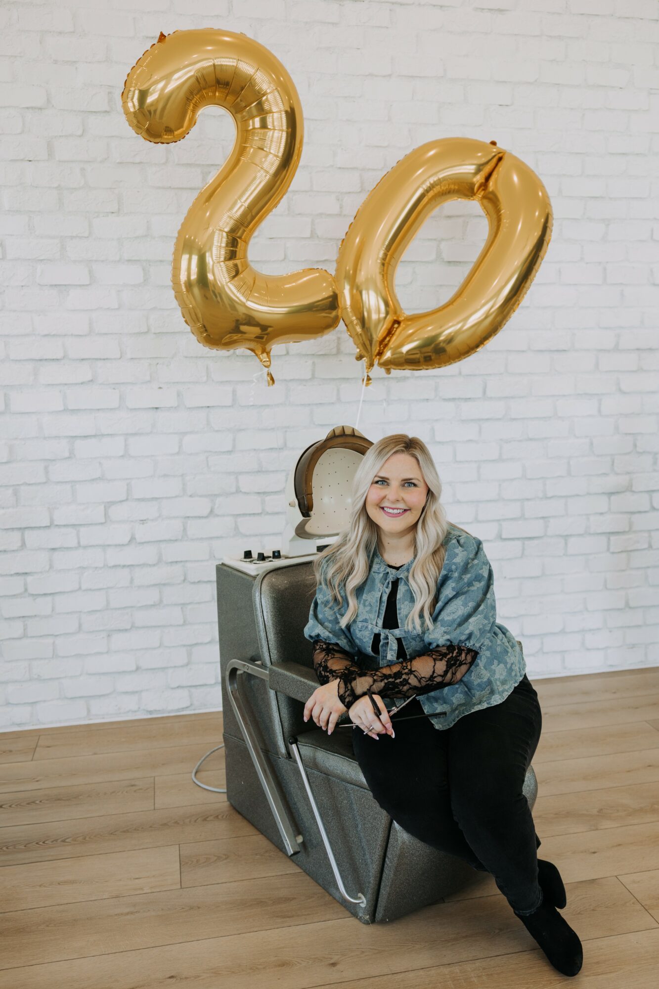 Woman sitting on a vintage suitcase with a large gold '20' balloon above her, against a white brick wall.