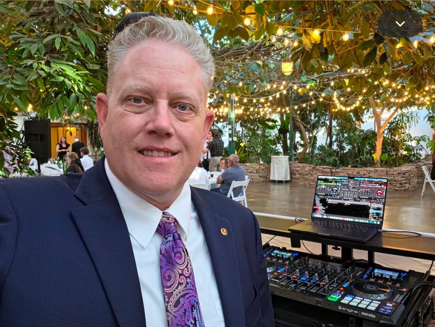 Man in suit and tie smiling outdoors with string lights and trees in background.