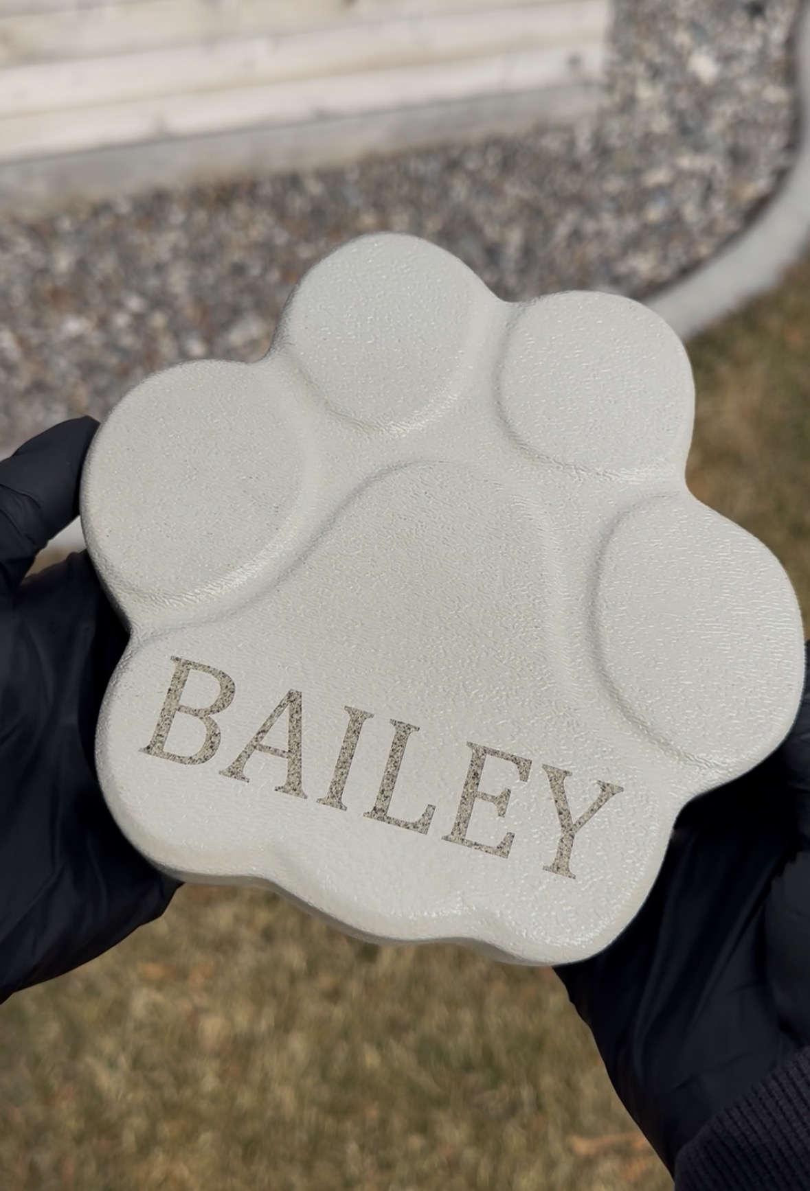Person holding a white paw-shaped object with the word BAILEY engraved, outdoors with grass and gravel background.