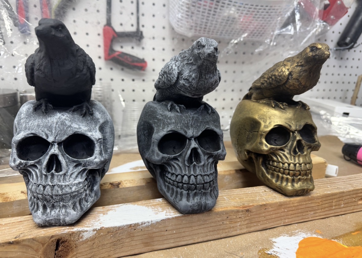 Three skull decorations with bird figures on top, in black, gray, and gold colors, placed on a wooden surface.
