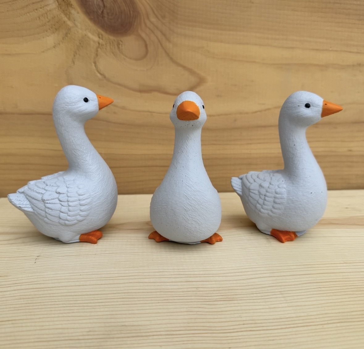 Three white duck figurines with orange beaks and feet, standing on a wooden surface against a wooden background.