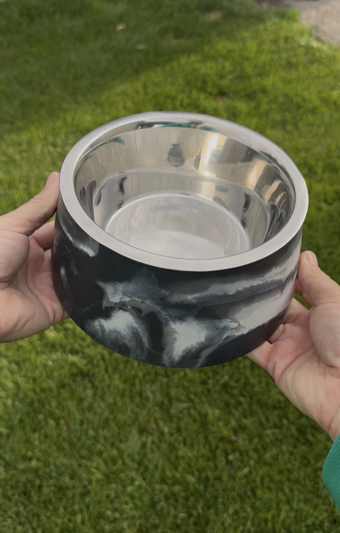 Person holding a round, reflective metal bowl outdoors on green grass, with hands visible on both sides.