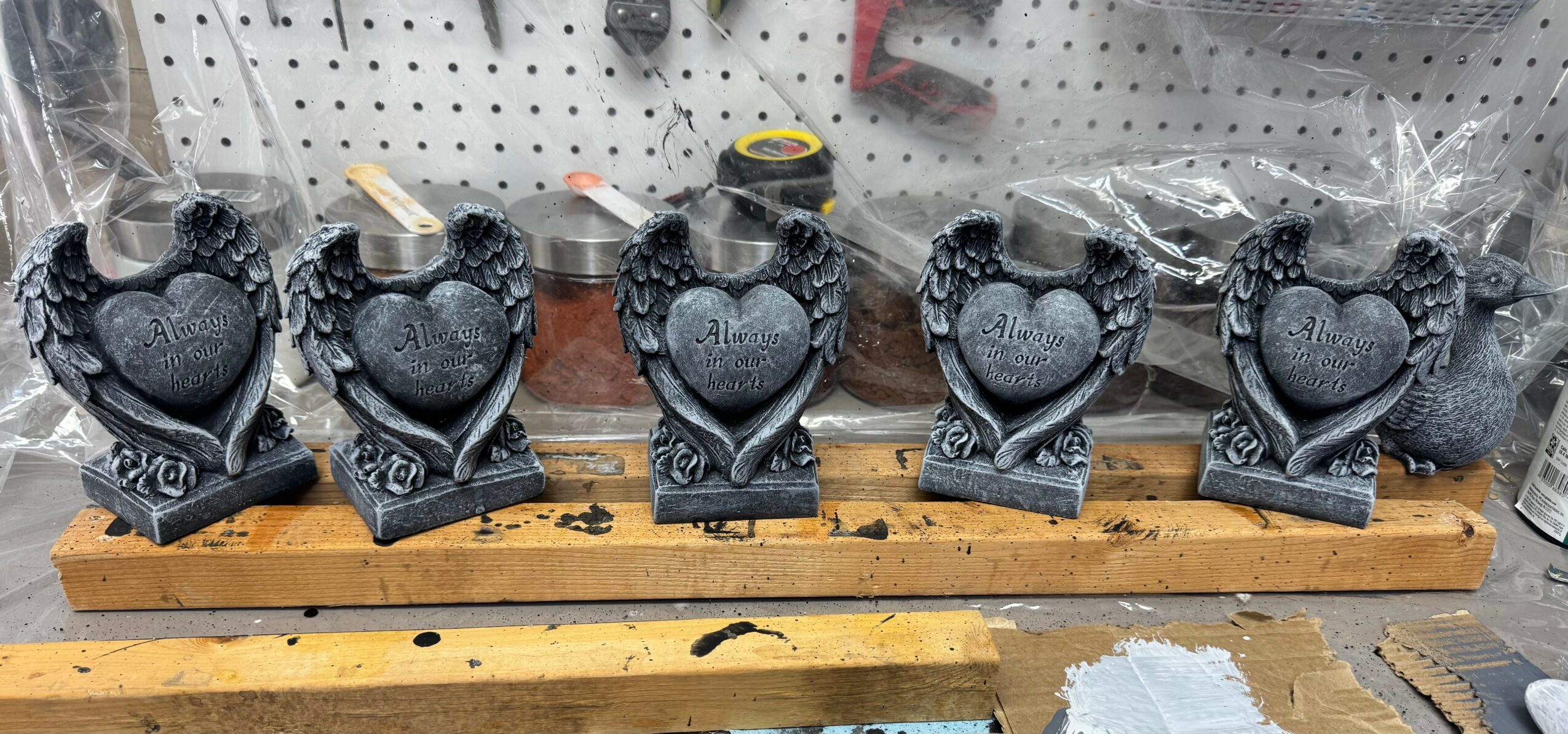 Five black heart-shaped candles with decorative bases, wrapped in plastic, placed on a wooden shelf in a workshop setting.