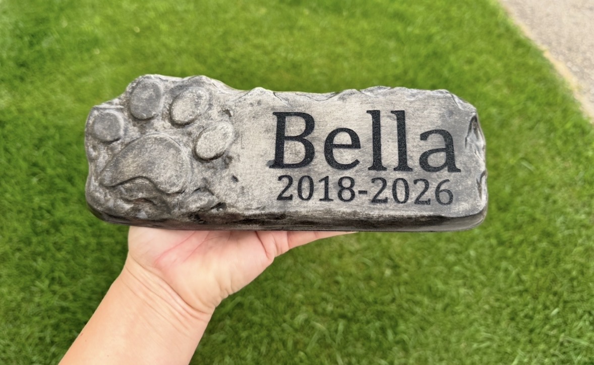Hand holding a stone with paw prints and engraved text, on green grass background.