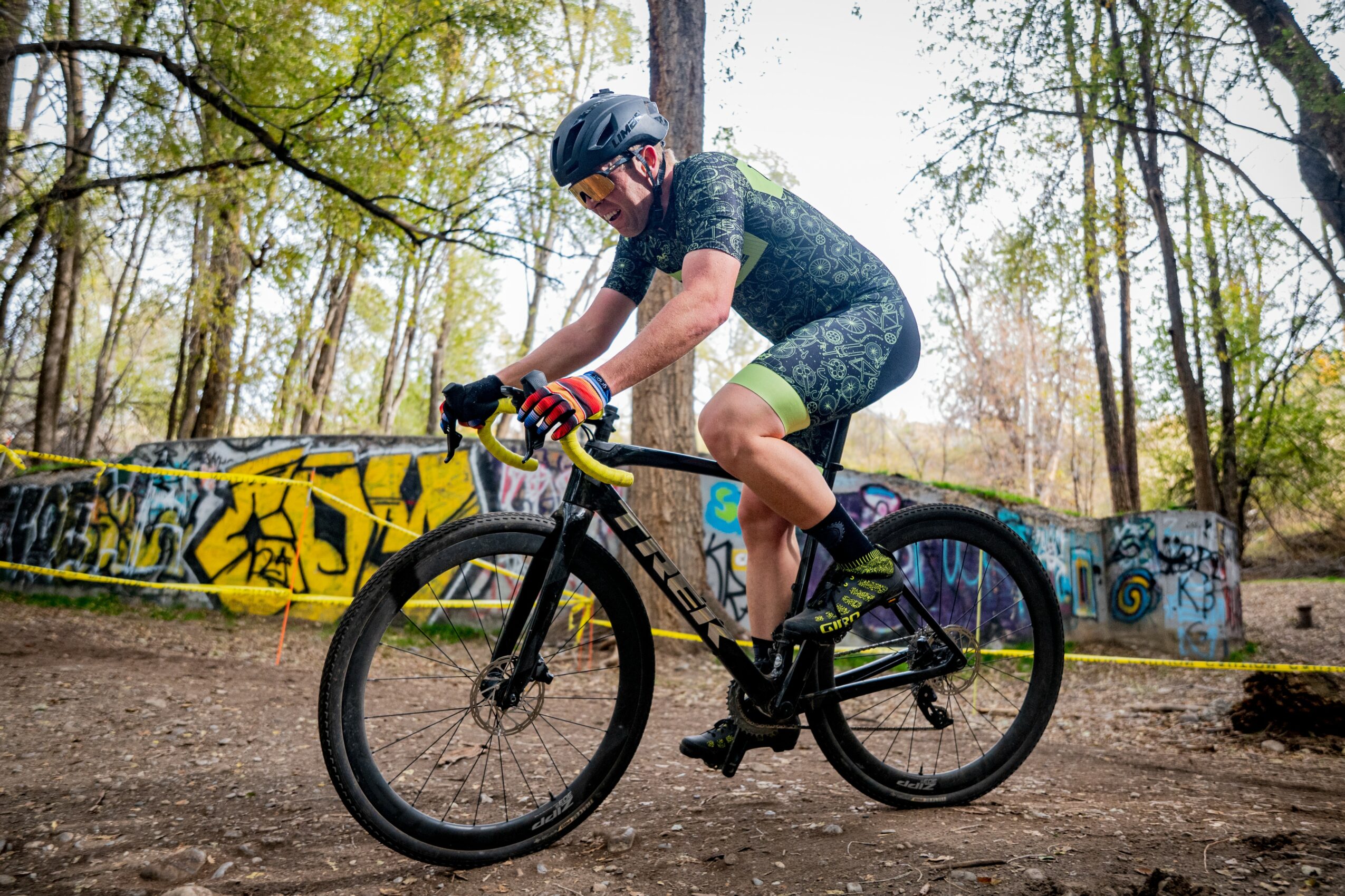 Person riding a mountain bike on a dirt trail in a wooded area, wearing a helmet and cycling gear.