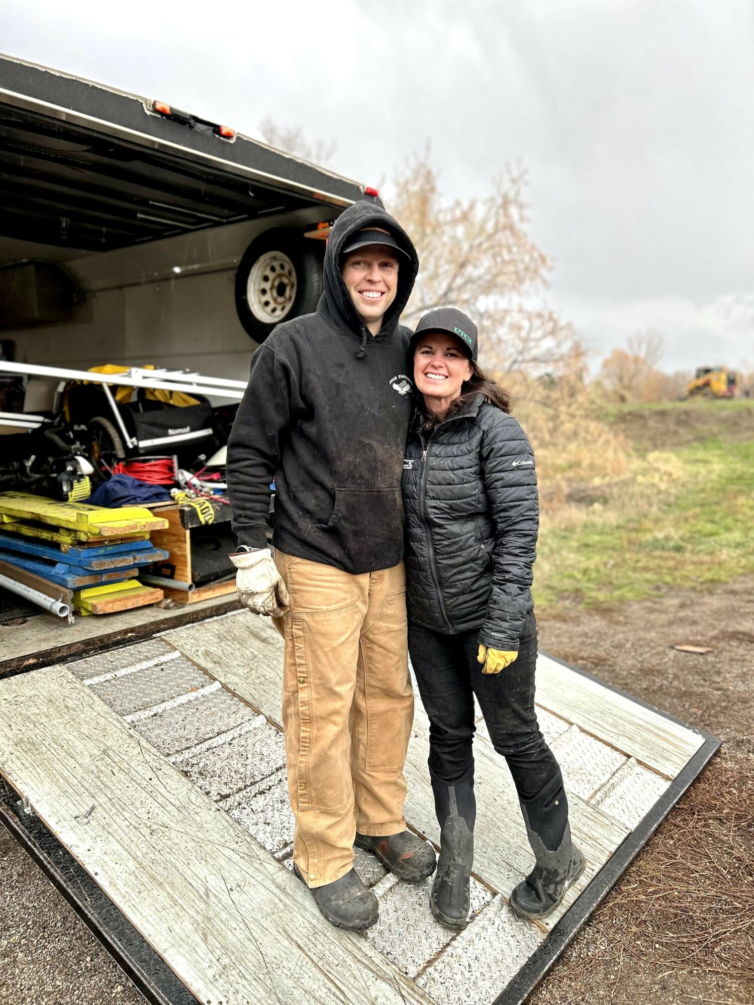 Two people wearing helmets and jackets stand on a ramp outdoors, smiling, with a trailer and equipment in the background.