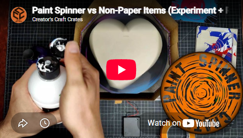 Hand holding a paint spinner with a container and a circular orange item on a grid surface.