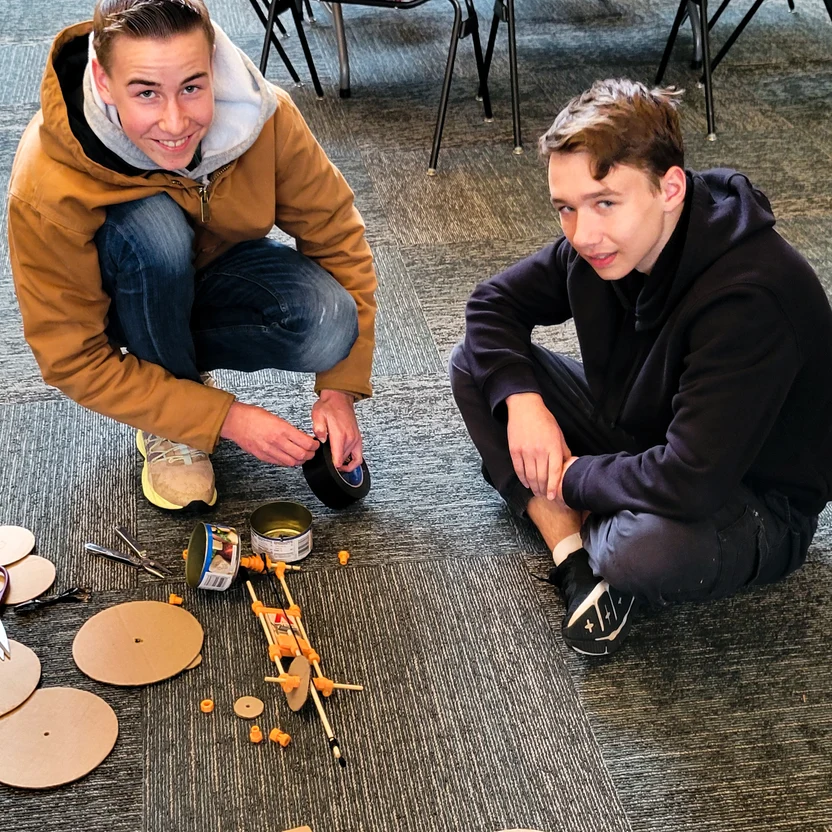 Two young people sitting on a carpeted floor with craft supplies and wooden circles, smiling at the camera.