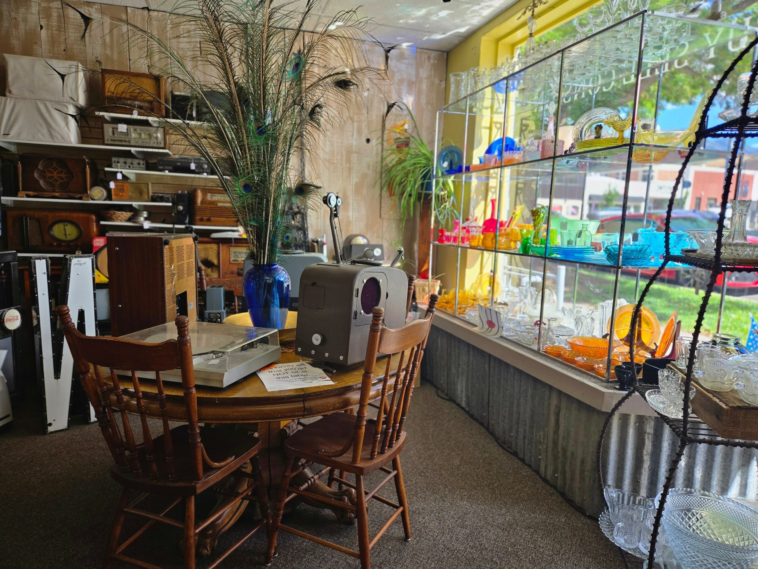 Interior of a cozy cafe with a round wooden table, four chairs, a large window with colorful decorations, and shelves with various items.