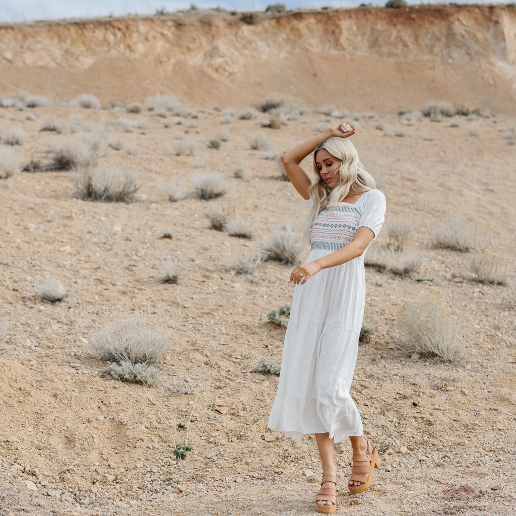 Woman in white dress standing in desert landscape with sparse vegetation, hand on head, looking down, wearing sandals.
