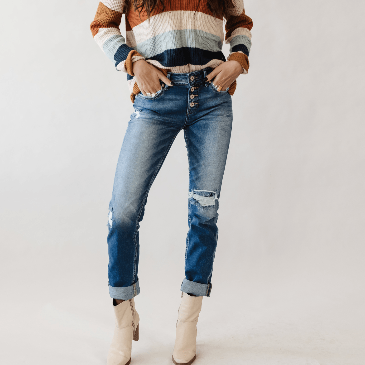 Person wearing a striped sweater, ripped jeans, and beige boots, standing with hands in pockets against a plain background.