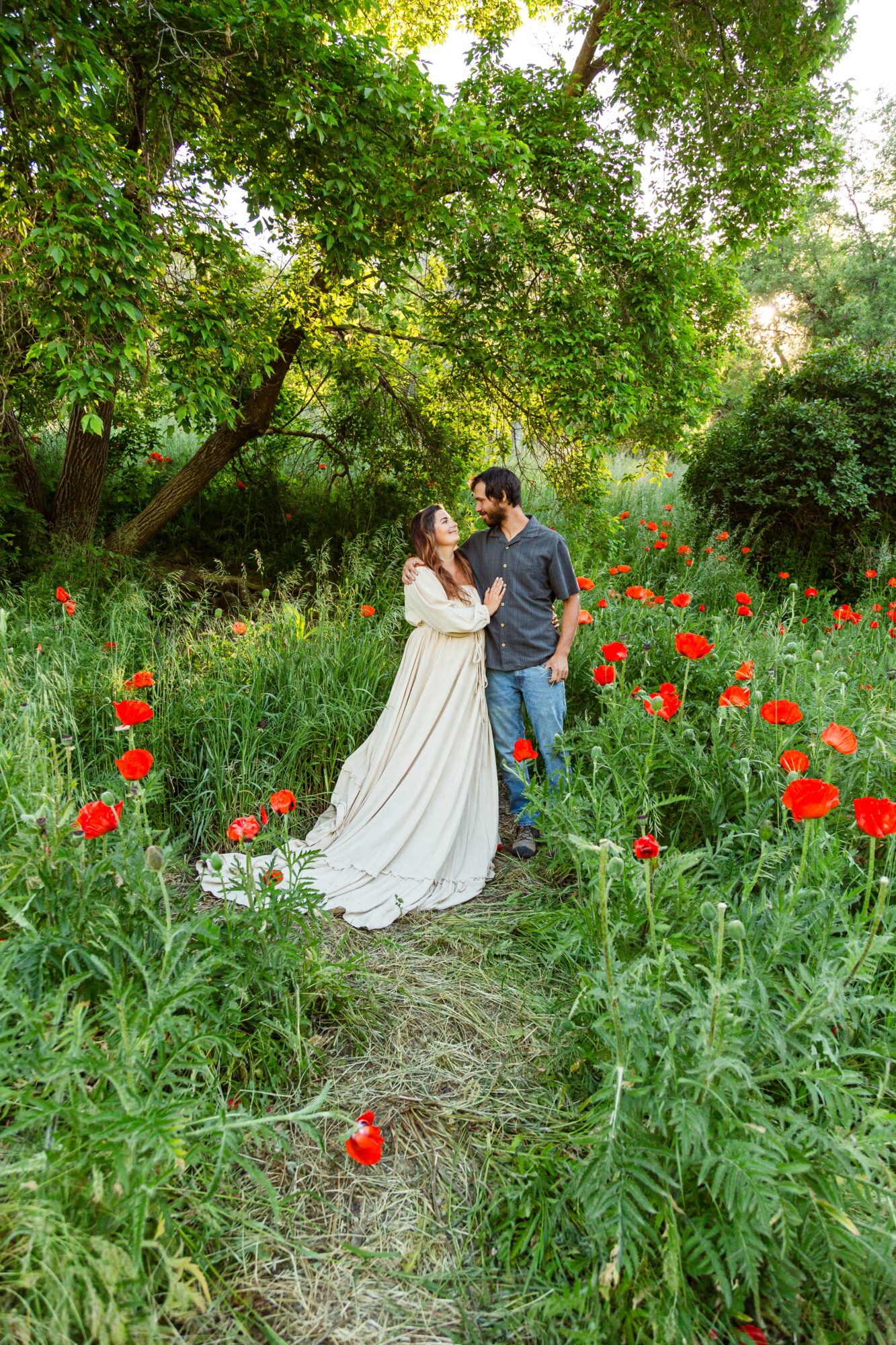 Couple standing on a garden path surrounded by red flowers and green trees, holding hands and looking at each other.