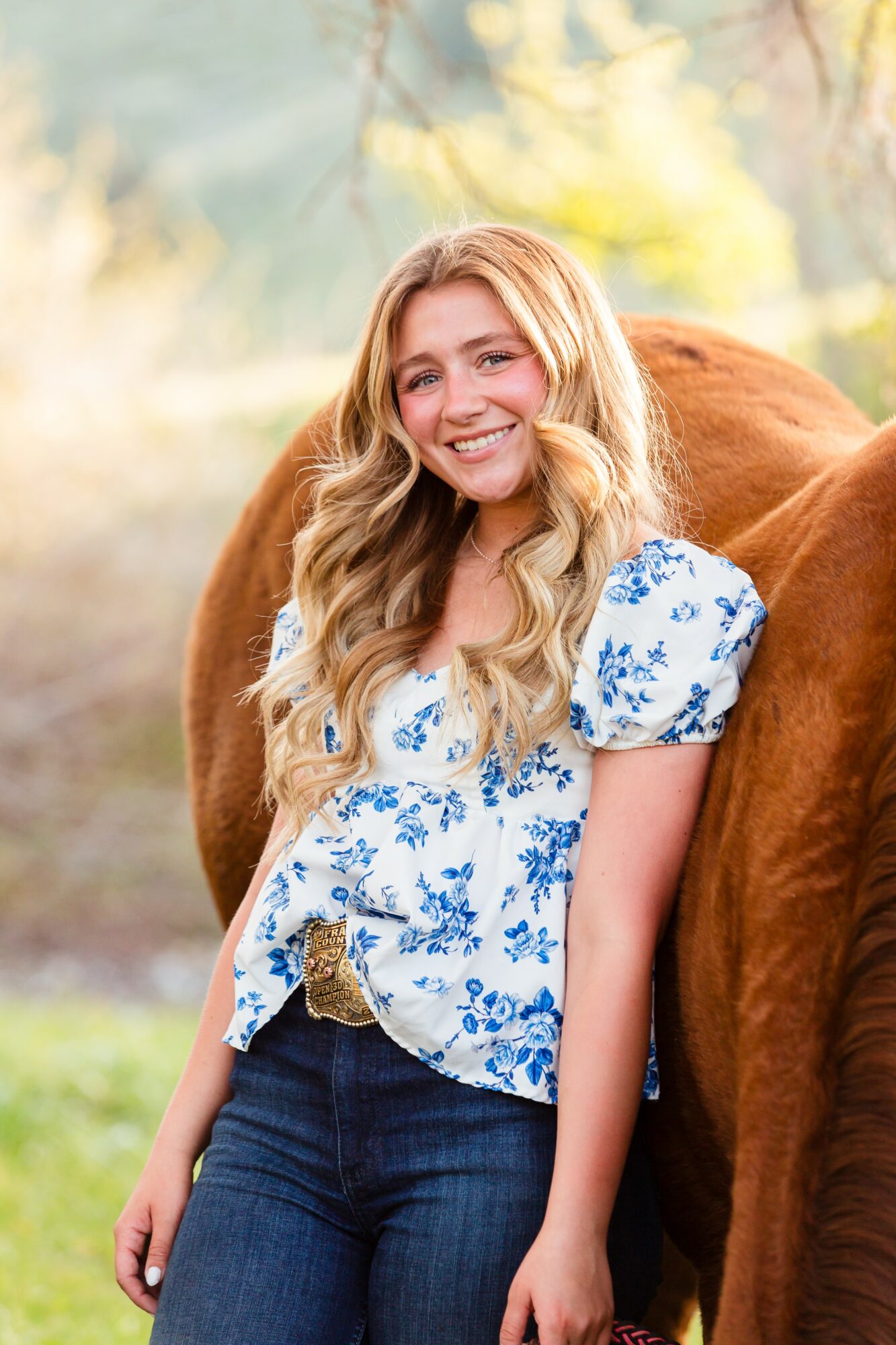 Young woman with long wavy blonde hair smiling outdoors, wearing a white floral blouse and jeans, standing next to a brown horse.