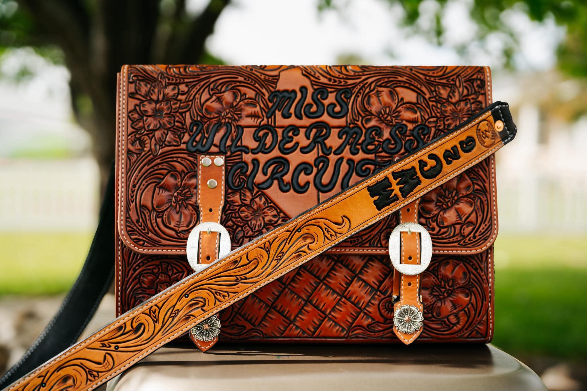 Decorative leather bag with intricate carved patterns and a strap with the word 'JUNG' on it, outdoors with trees in background.