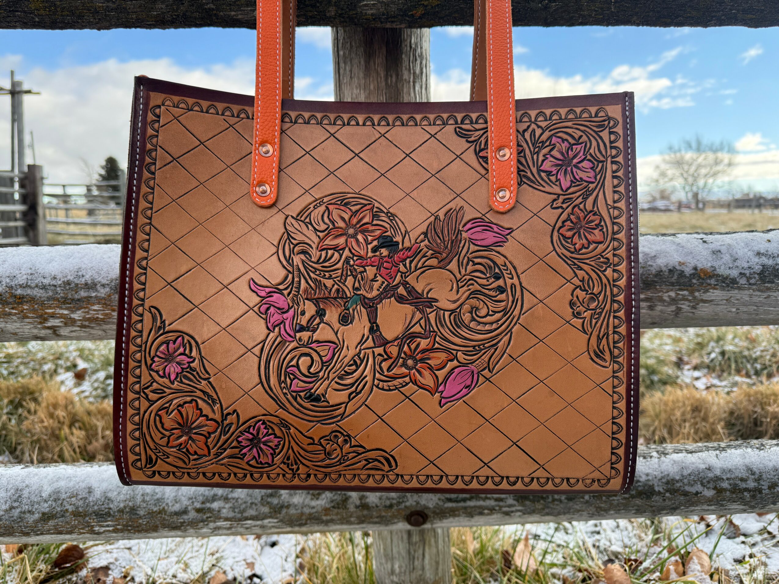 Leather handbag with floral and animal designs, orange handles, hanging outdoors with a snowy landscape and blue sky.