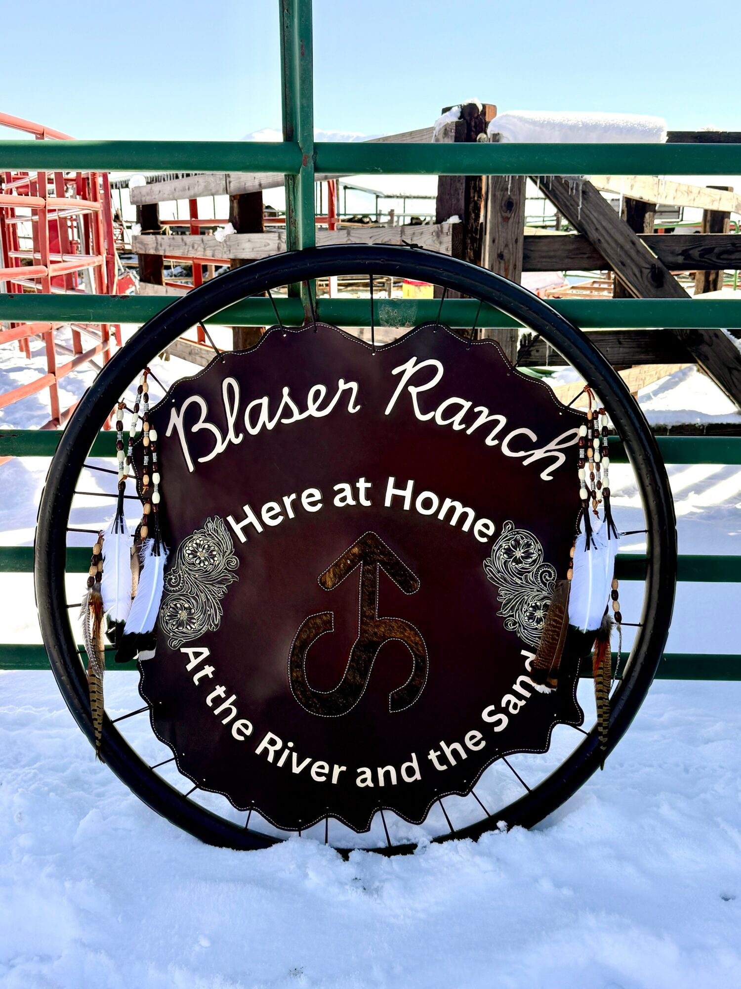 Round sign with text and a horse illustration, mounted on a green fence at a ranch, snow on the ground.