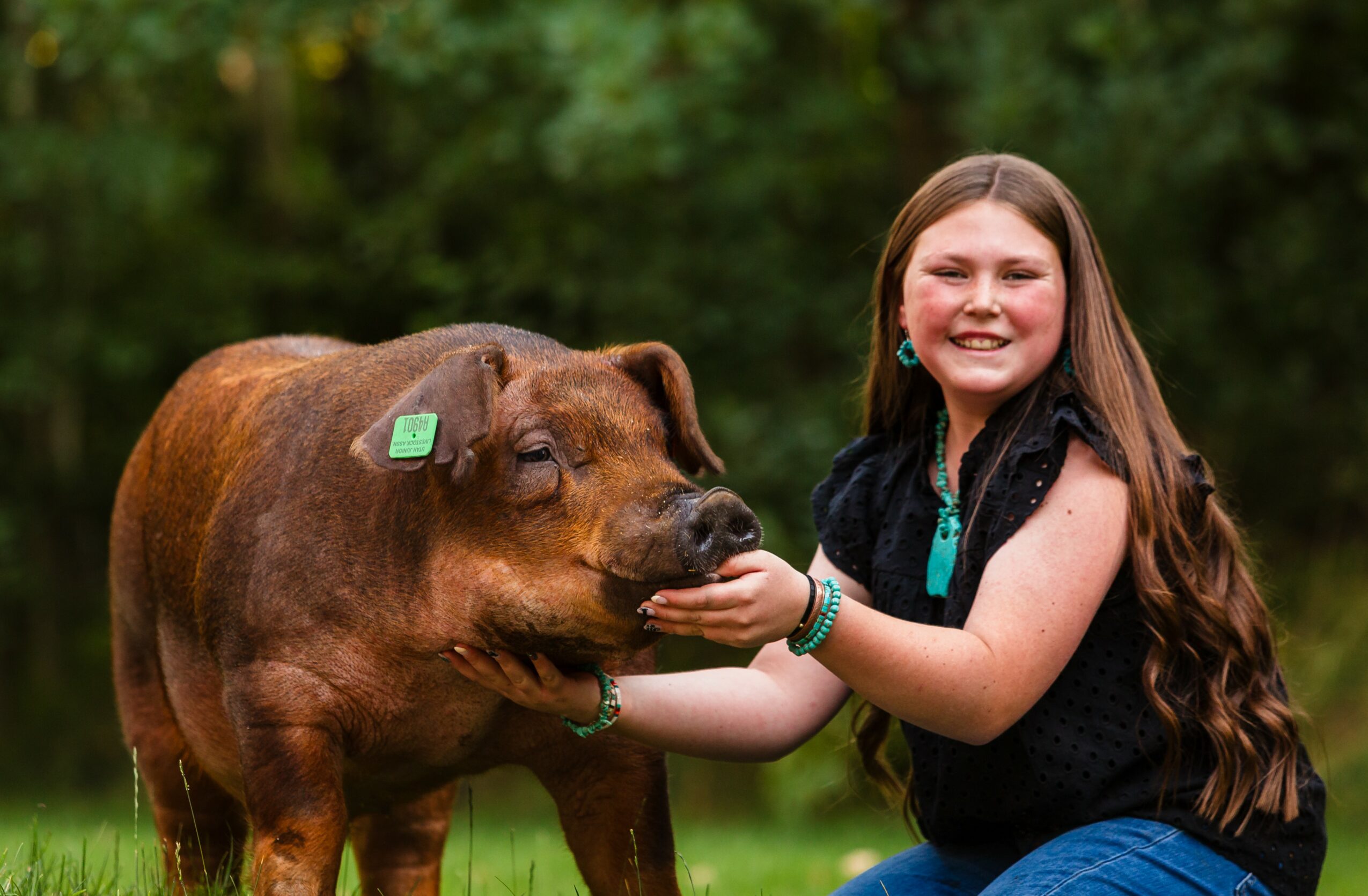 Girl smiling with a brown pig outdoors, holding its snout, green trees in background.