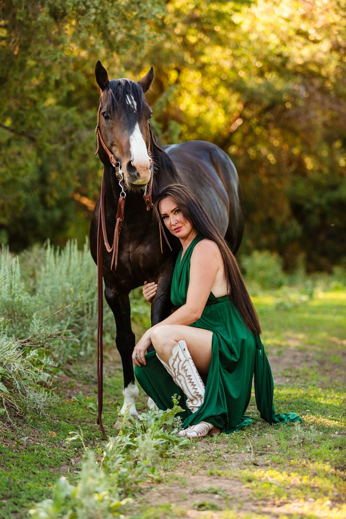 Woman in green dress kneels beside a black horse in a park with autumn trees in background.