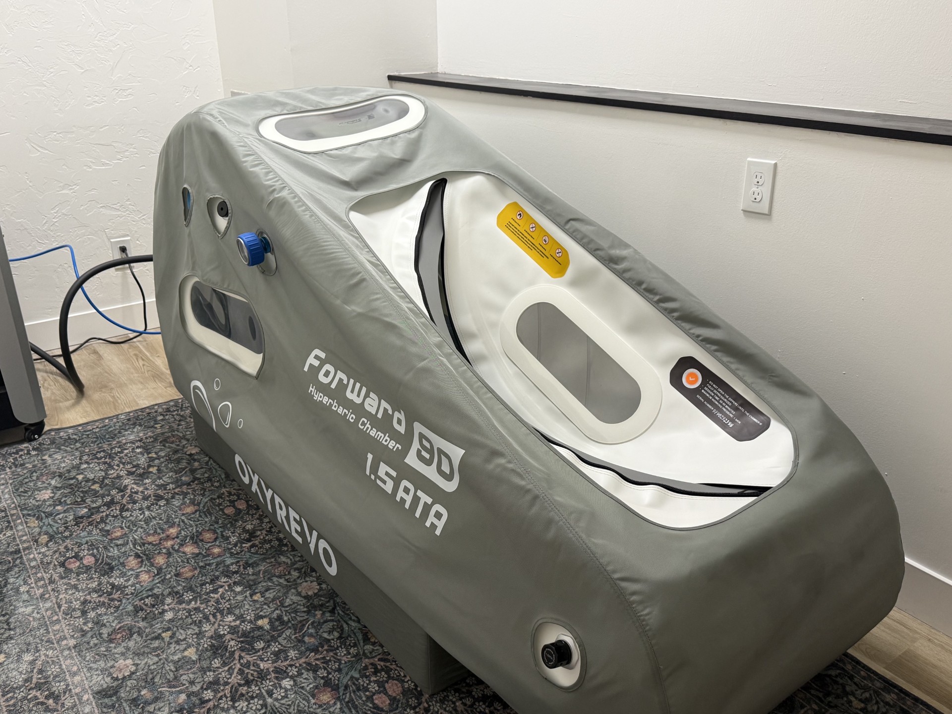 Medical hyperbaric chamber in a room with a white wall and electrical outlet, gray exterior with control panel and viewing window.