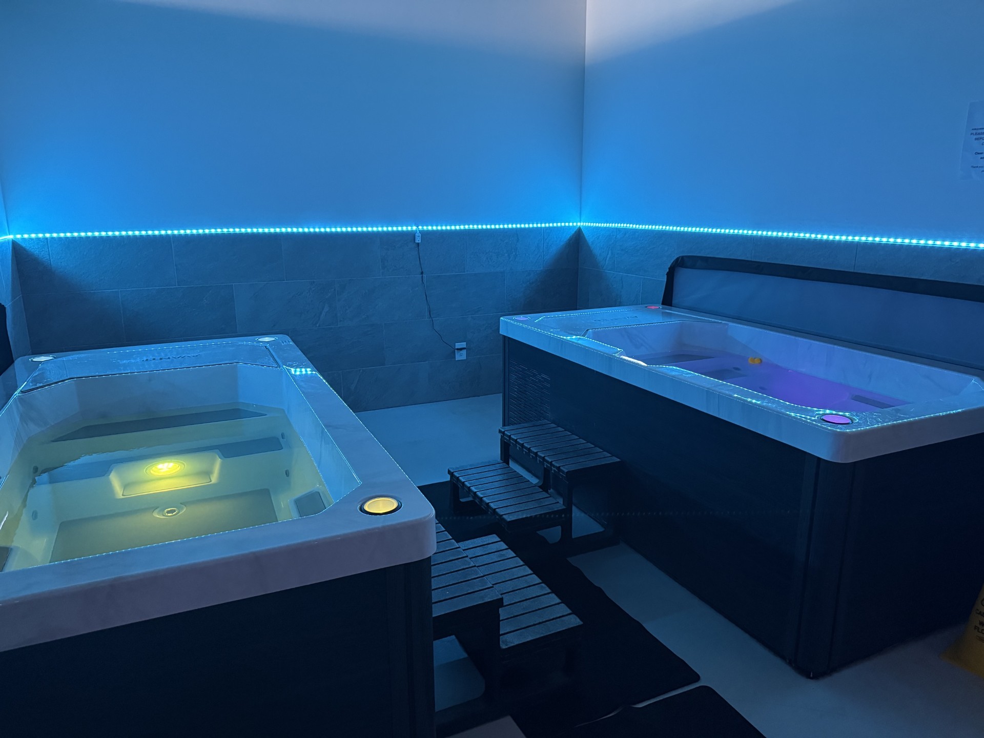 Two hot tubs in a room with blue lighting, one with a yellow light and the other with purple, connected by a small bridge.