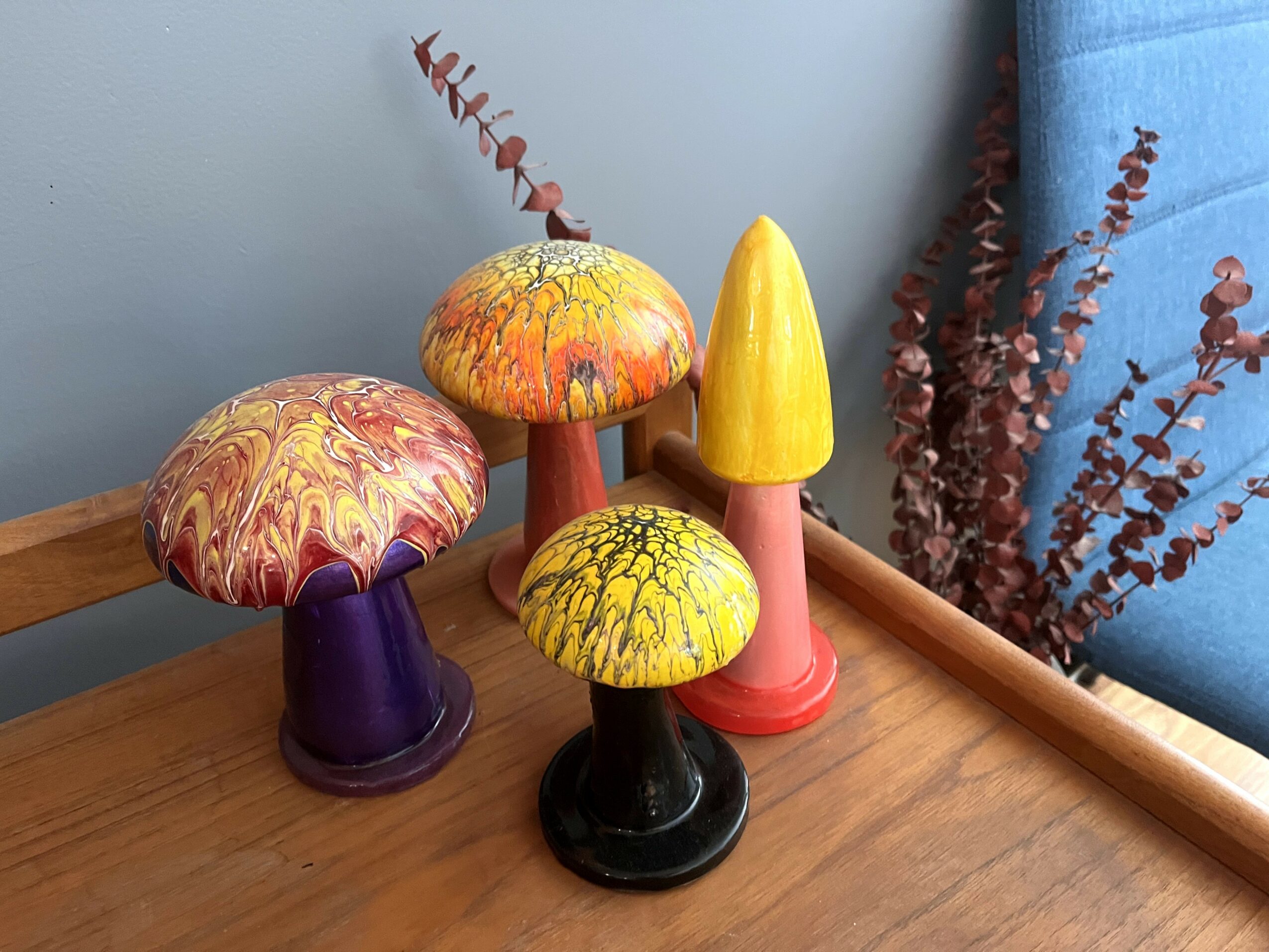 Four colorful mushroom-shaped decorations on a wooden surface near a blue curtain and dried plants.