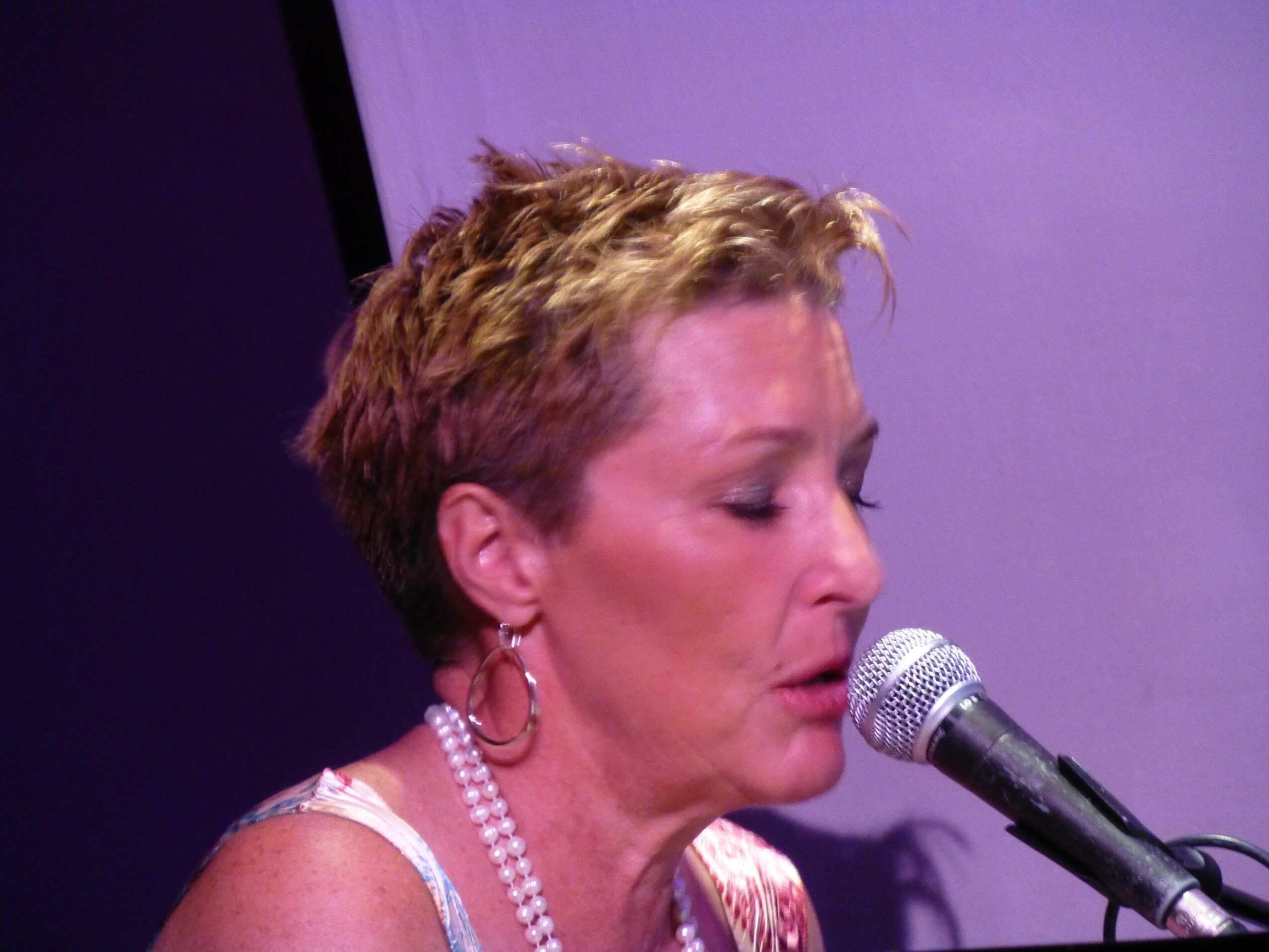 Woman with short hair singing into a microphone, wearing earrings and a pearl necklace.