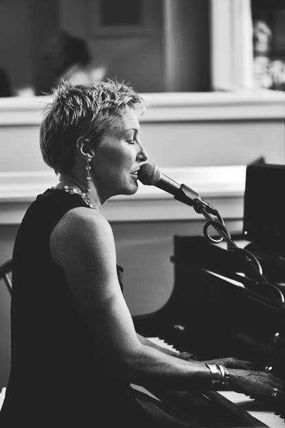 Woman with short hair singing into a microphone while playing a piano, in black and white.