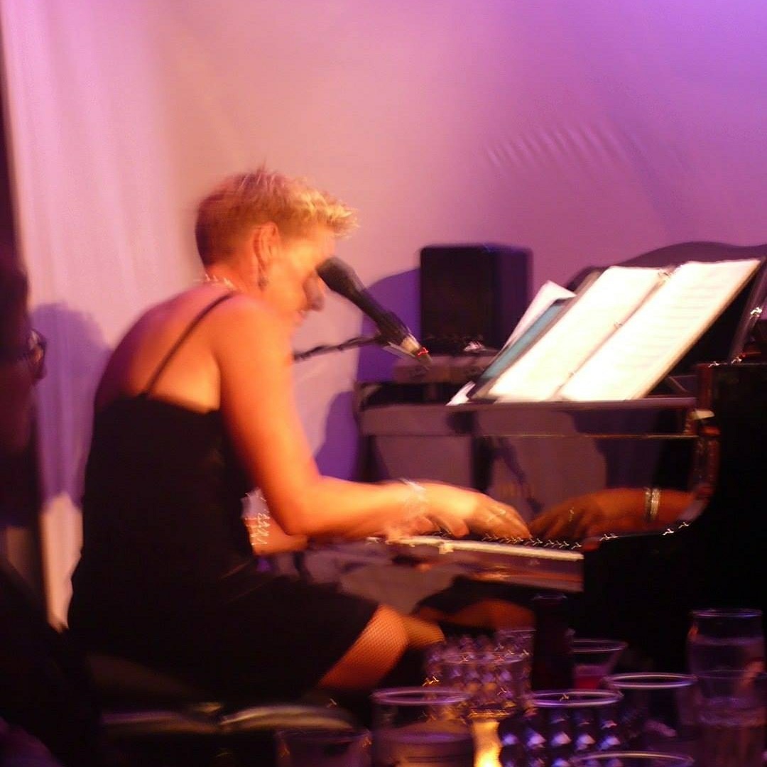 Person playing piano with sheet music, in a dimly lit room with purple lighting, wearing a black dress.