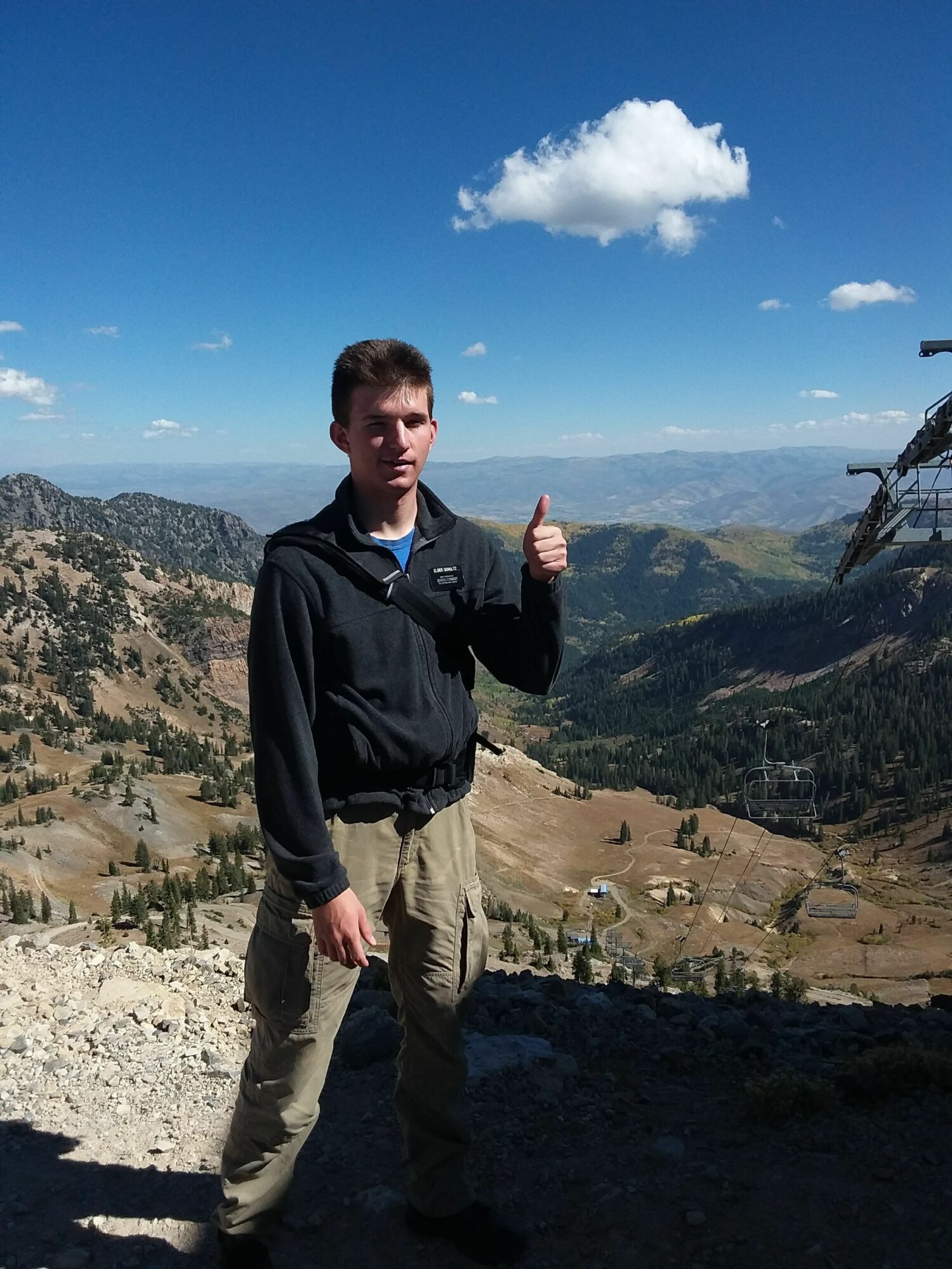 Young man standing outdoors with mountains and blue sky in background.