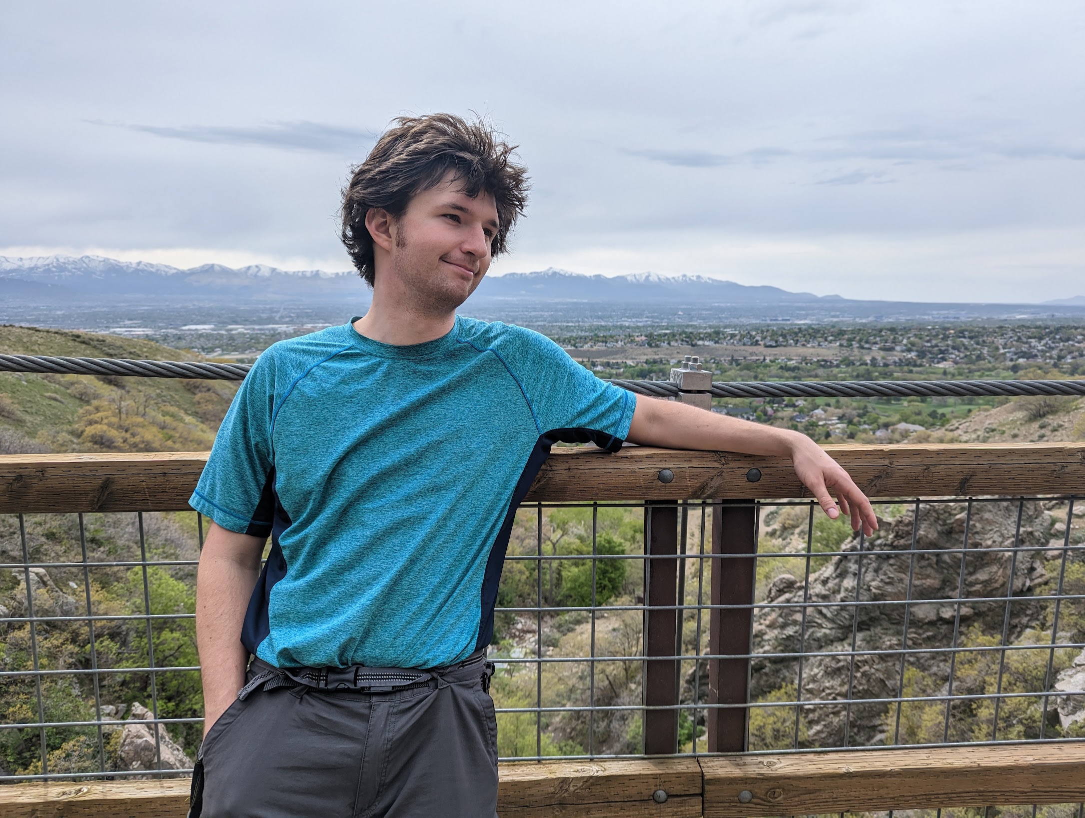 Young person with curly hair leaning on a wooden railing outdoors, with a landscape of trees and sky in the background.