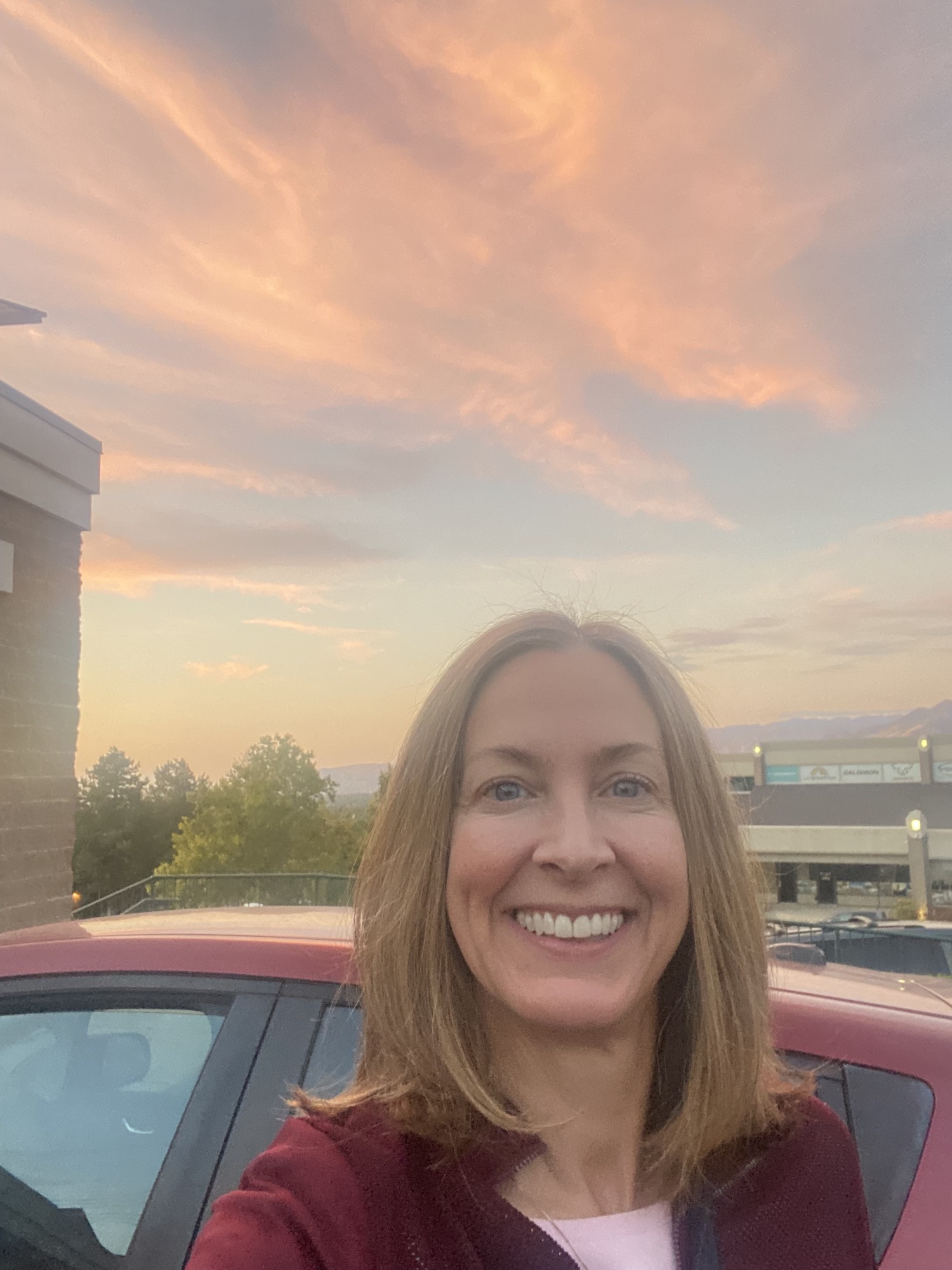 Smiling woman with shoulder-length hair outdoors during sunset, with a car and building in the background.
