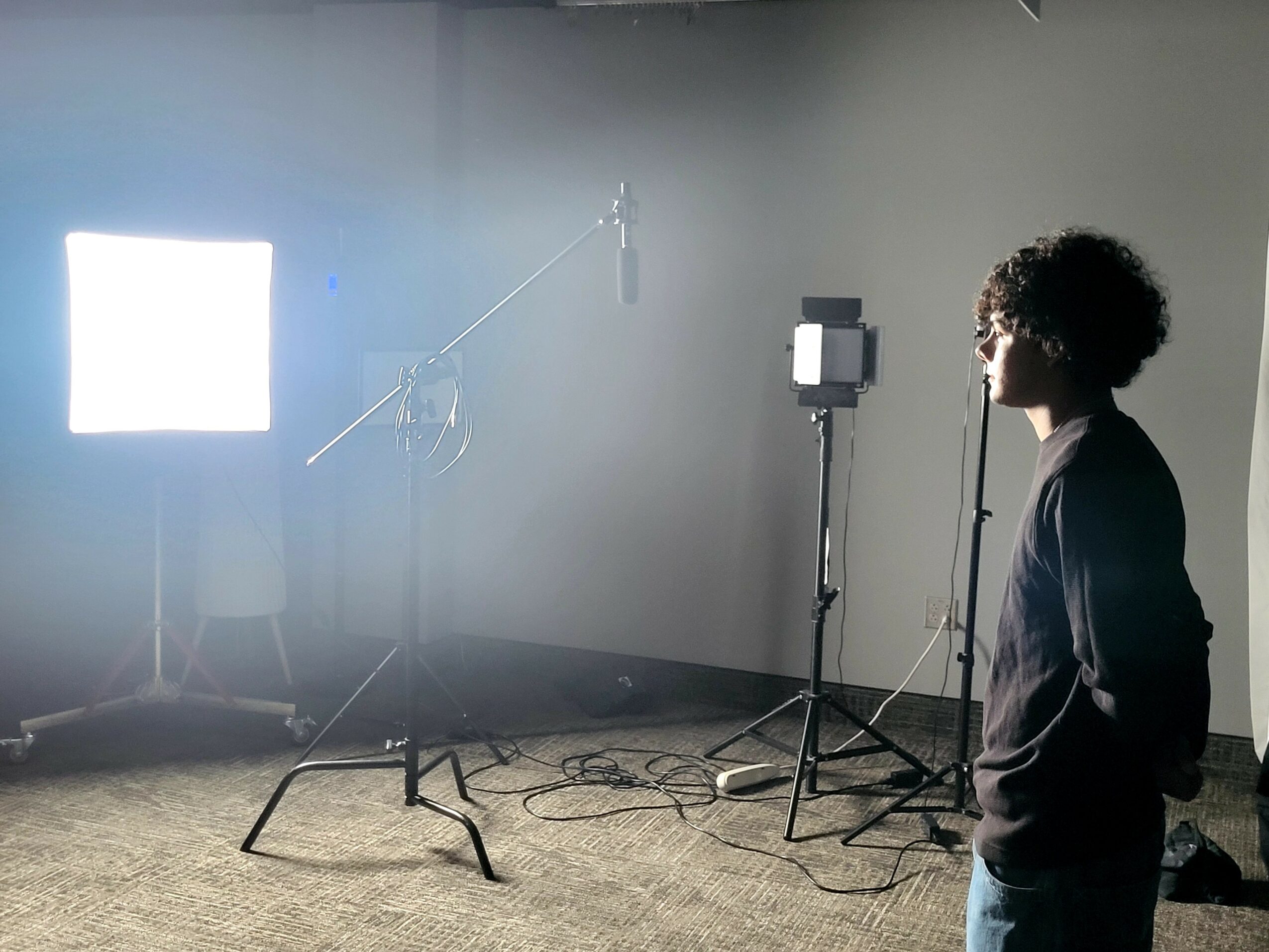 Person with curly hair stands in a studio with lighting and equipment, facing a bright light source on the left.