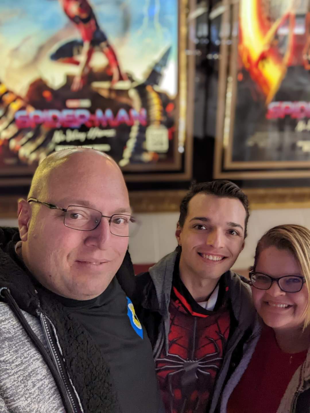 Three people smiling in front of Spider-Man movie posters, two men and one woman, indoors.