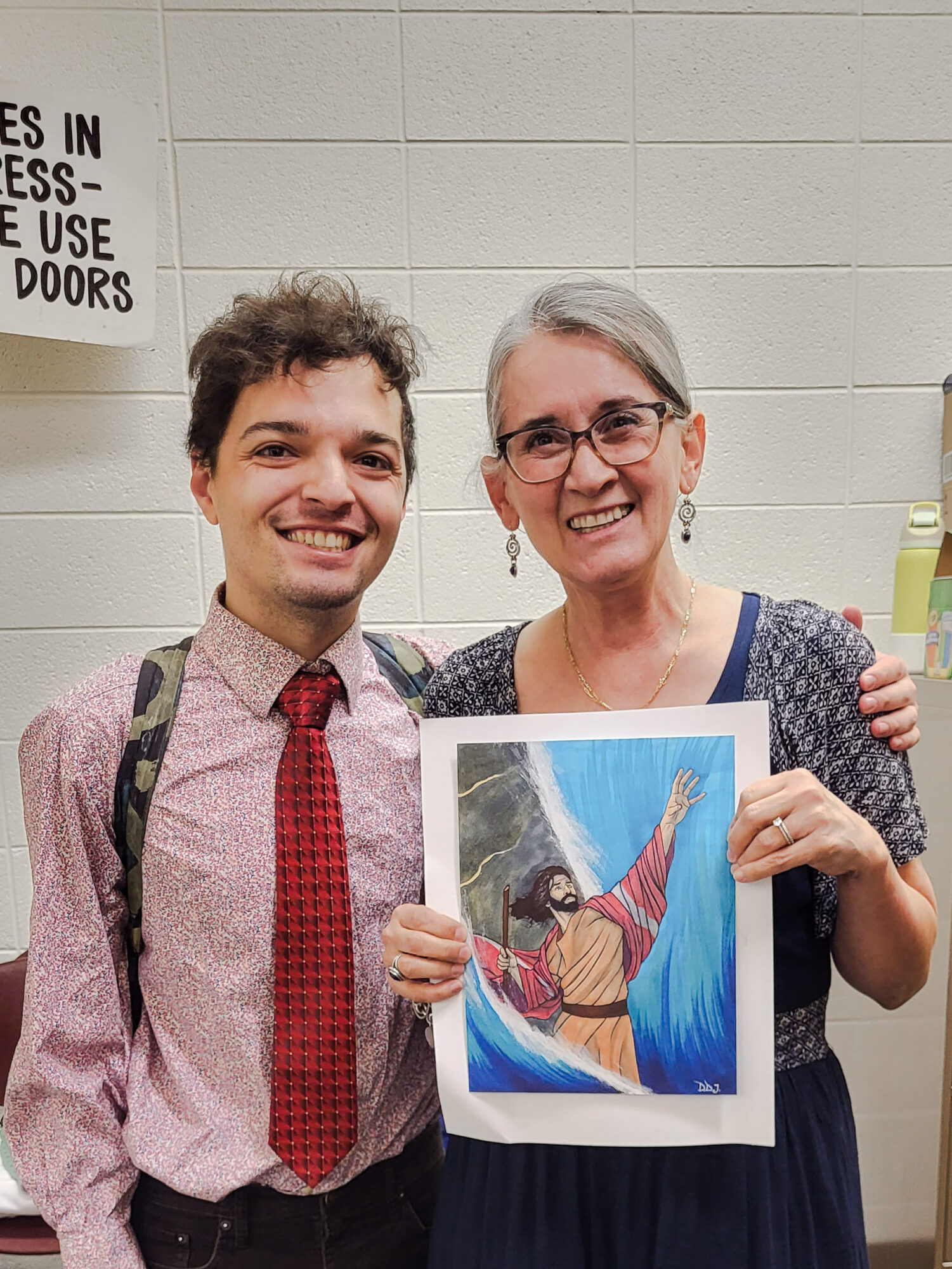 Two people smiling, one holding a colorful drawing of a woman with outstretched arm, in front of a white wall.