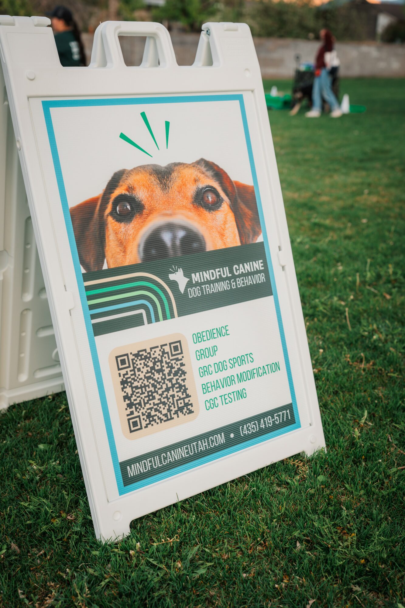 Signboard with a dog's face, QR code, and information about a canine event, placed on grass in a park.