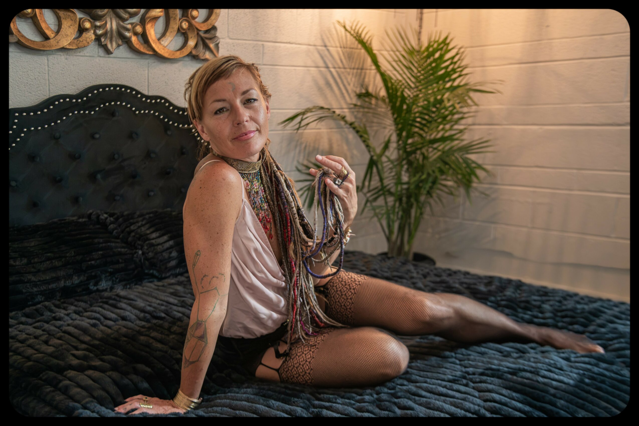 Woman sitting on a bed with a black headboard, holding a scarf, in a room with a plant and wooden wall panels.