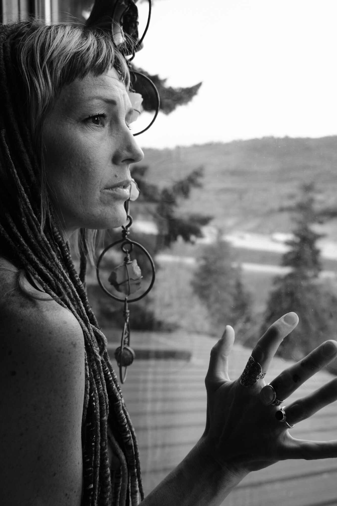 Side profile of a woman with dreadlocks and large earrings, outdoors with trees and landscape in background.