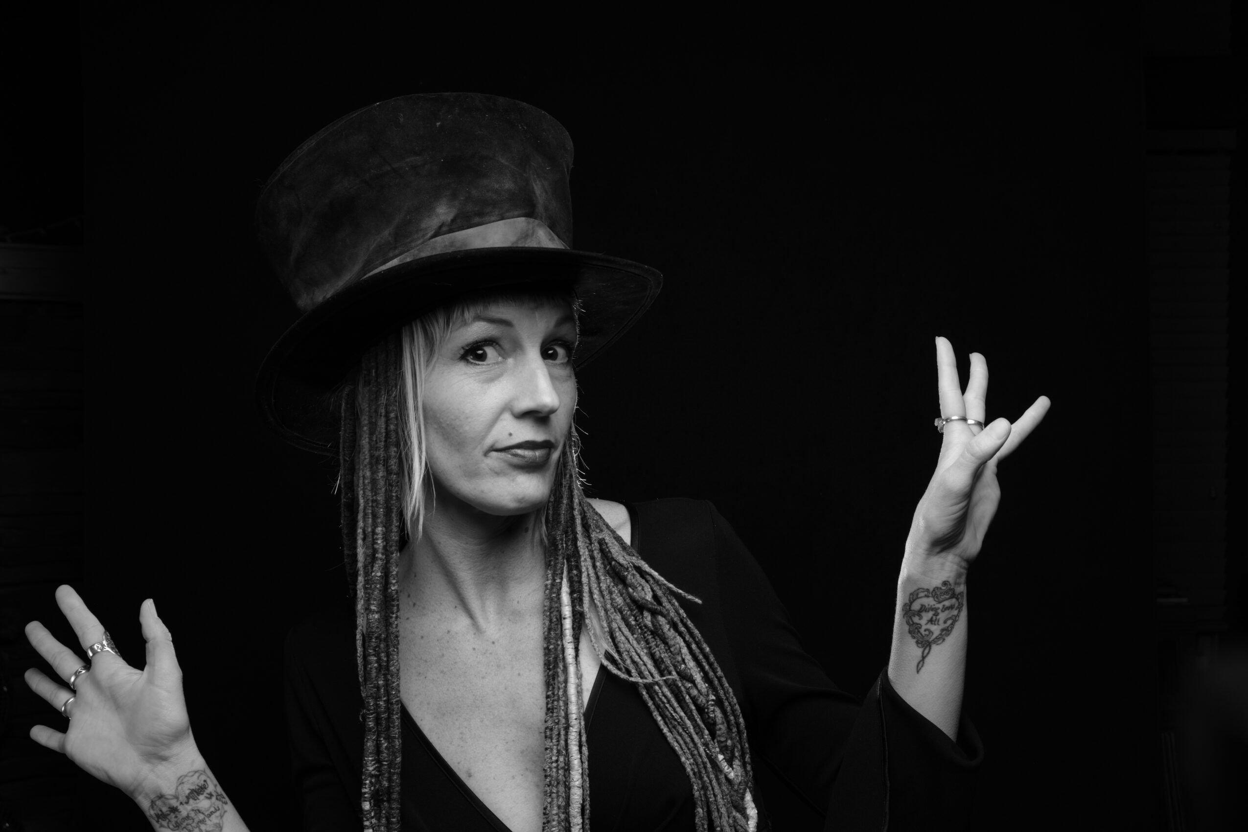 Woman wearing a top hat and with dreadlocks, making a gesture with her hands, in black and white.